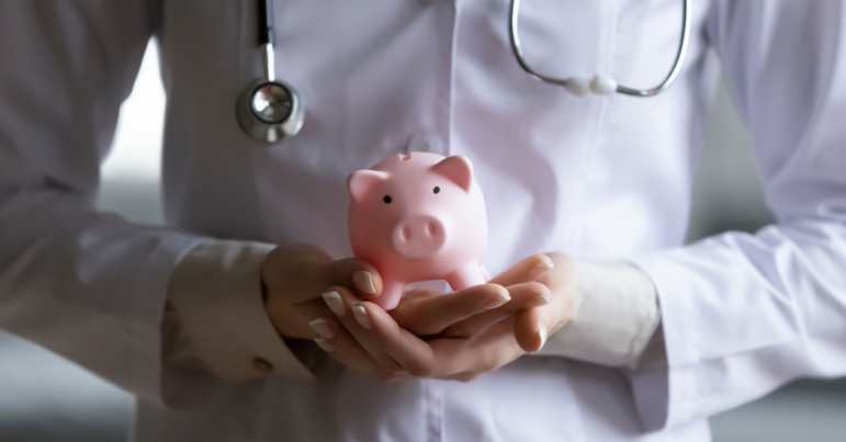 A physician's hands hold a piggy bank in front of their body